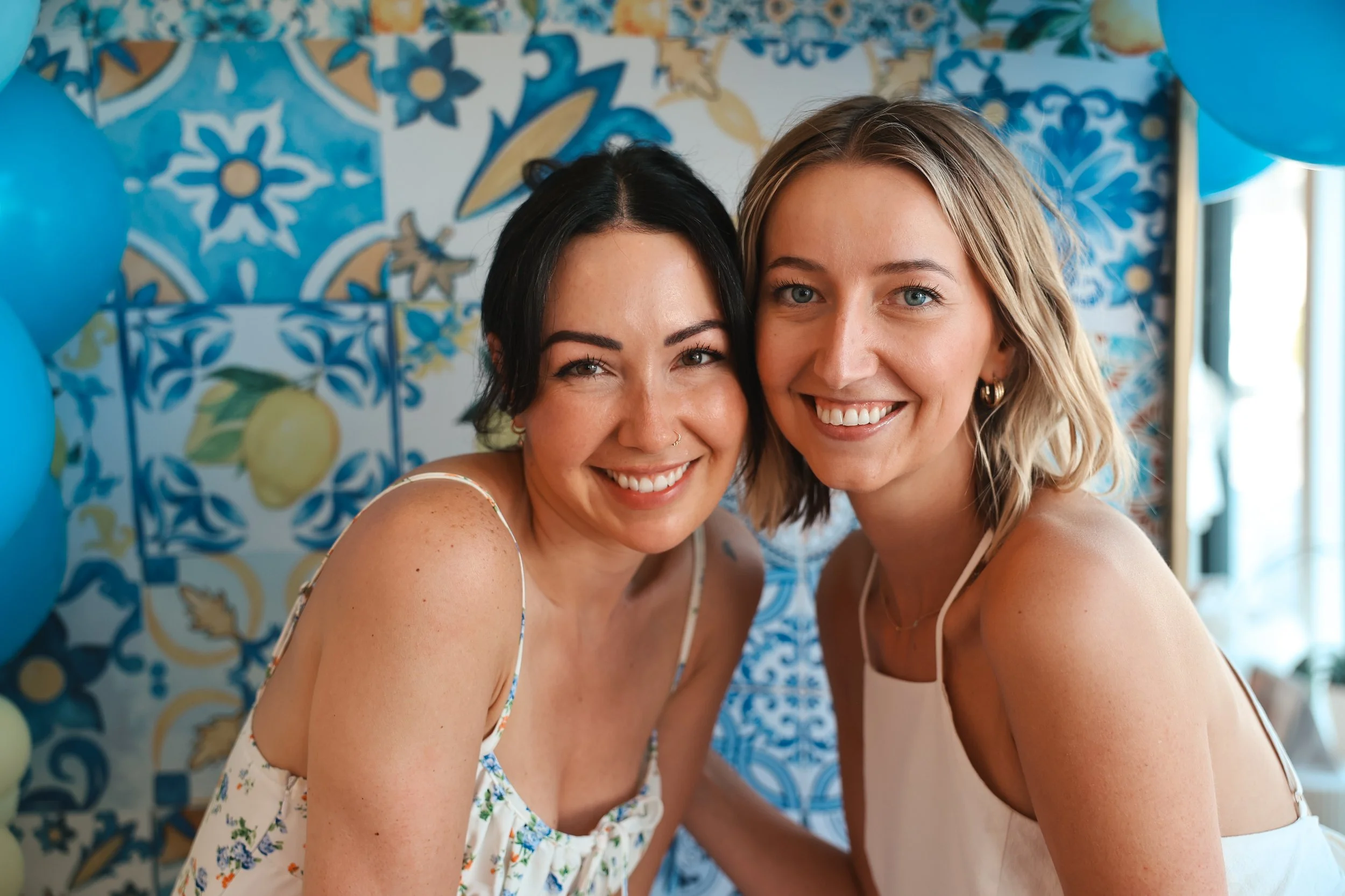 Two women smiling and posing at a party with blue balloons and patterned wallpaper with lemons and blue floral designs in the background.