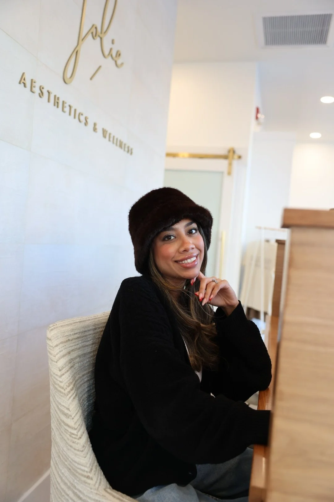 A woman wearing a black hat and black sweater is smiling while sitting at a counter in a wellness or aesthetics clinic.