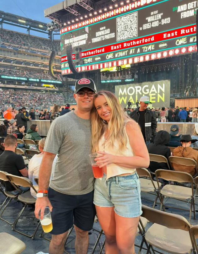 A young man and woman smiling and holding drinks at a concert or outdoor event, with a large stage and crowd in the background, and a digital display showing 'Morgan Wallen' and event details.