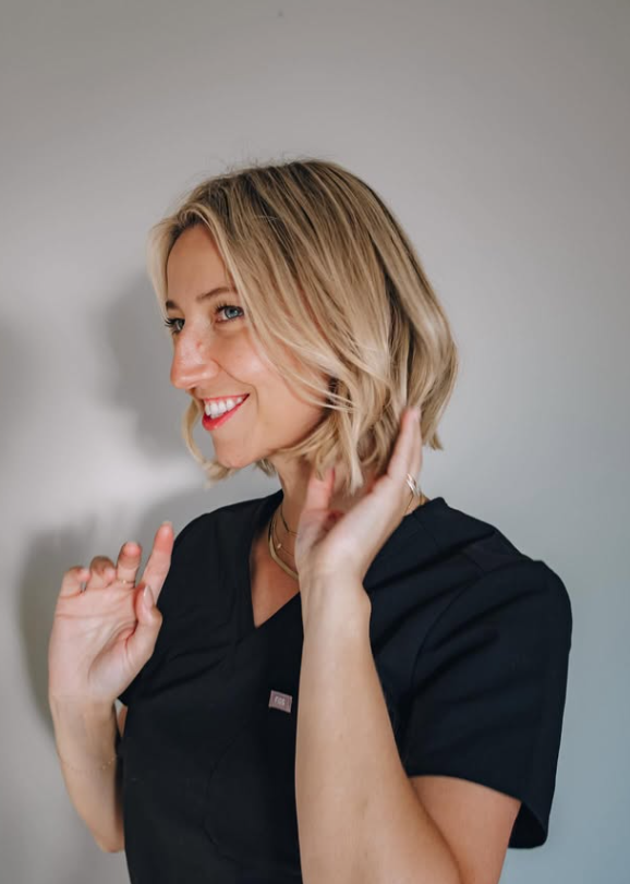 A smiling woman with blonde, shoulder-length hair, wearing a black uniform, posing against a plain gray background.