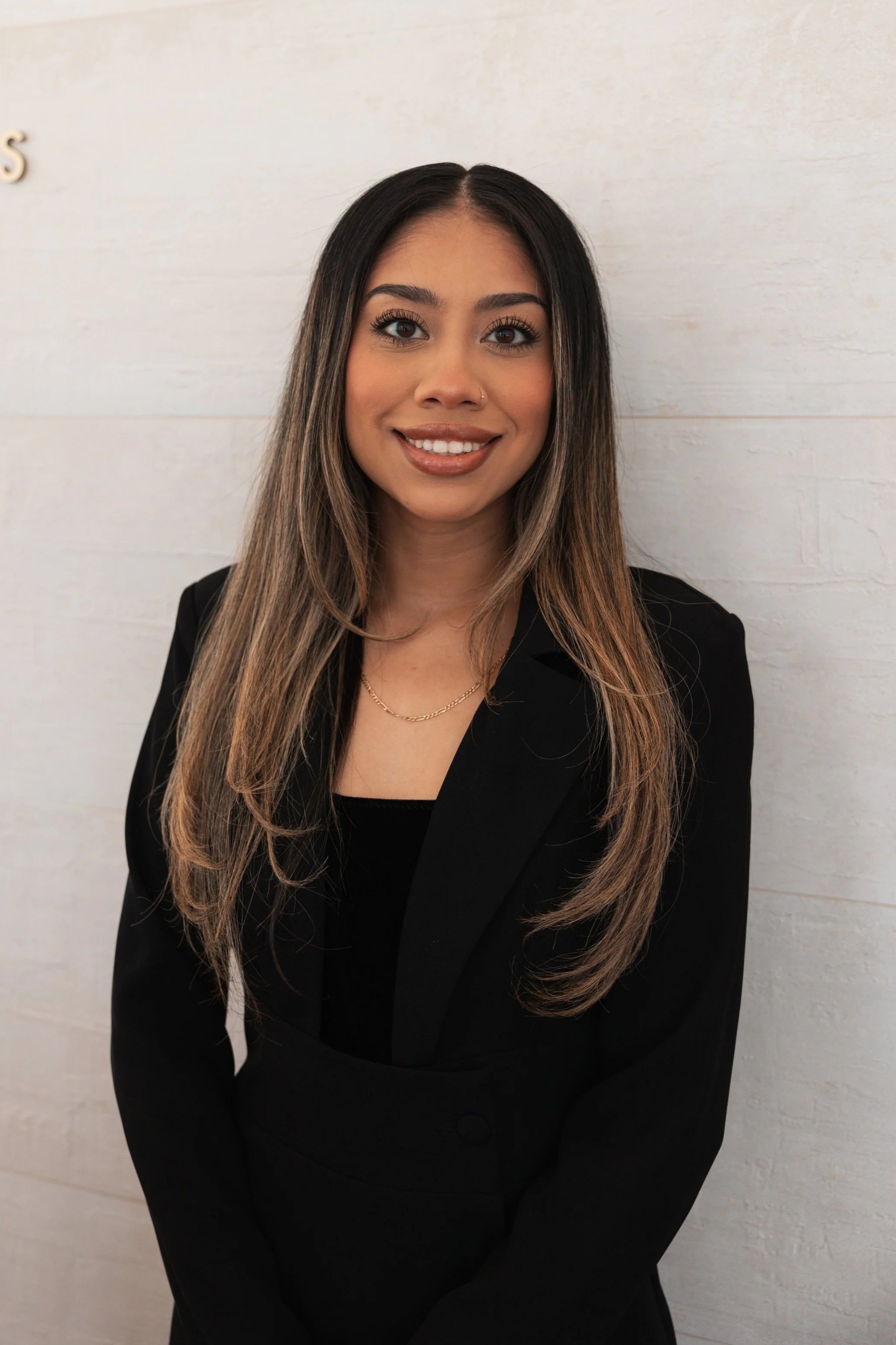 Young woman with long brown hair wearing a black blazer and a gold necklace, smiling and standing against a light-colored wall.