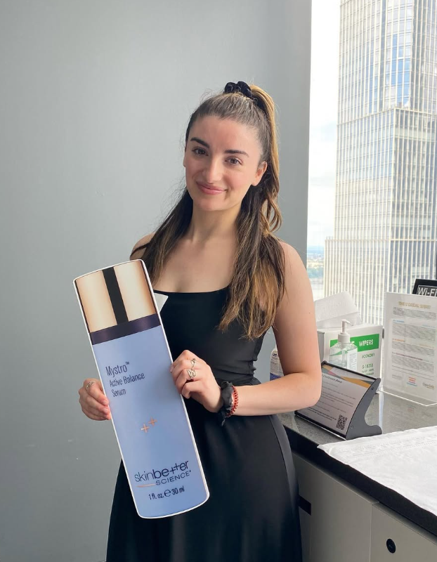 A young woman with long brown hair in a ponytail, wearing a black sleeveless dress, standing indoors near a window with a cityscape view, holding a large bottle of skincare product labeled 'Mystro Active Balance Serum'.