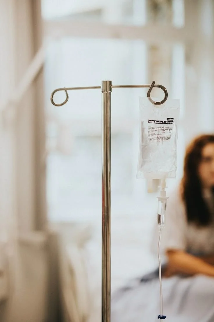 An IV drip stand with a bag of saline solution attached, hanging in a medical setting, with a blurred person sitting in the background.