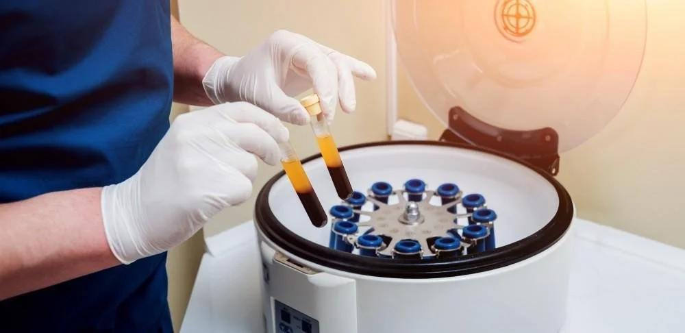 A scientist wearing white gloves handling test tubes with blood samples, placed inside a centrifuge machine in a laboratory setting.