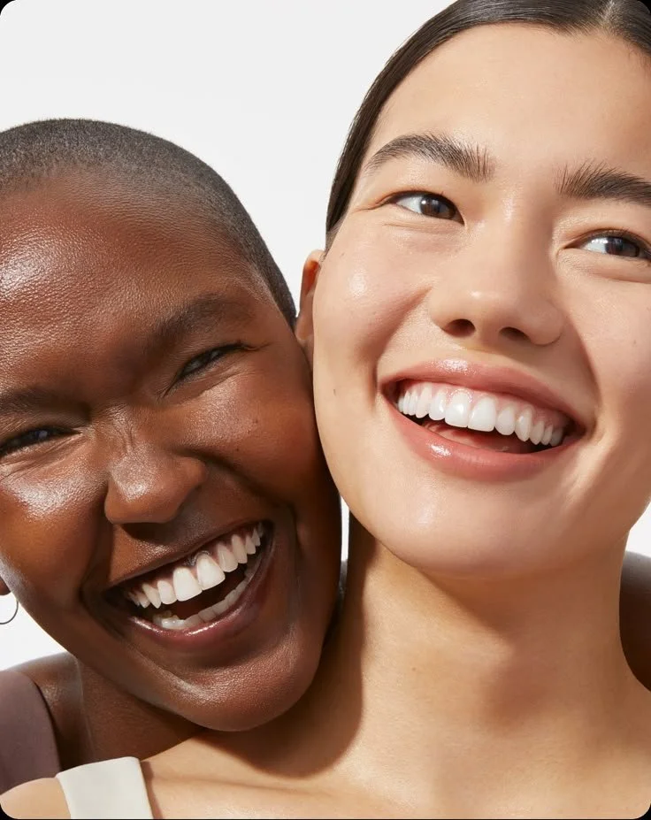 Close-up of two women smiling and showing teeth, with their faces close together. One woman has dark skin and short hair, and the other has light skin and long hair.