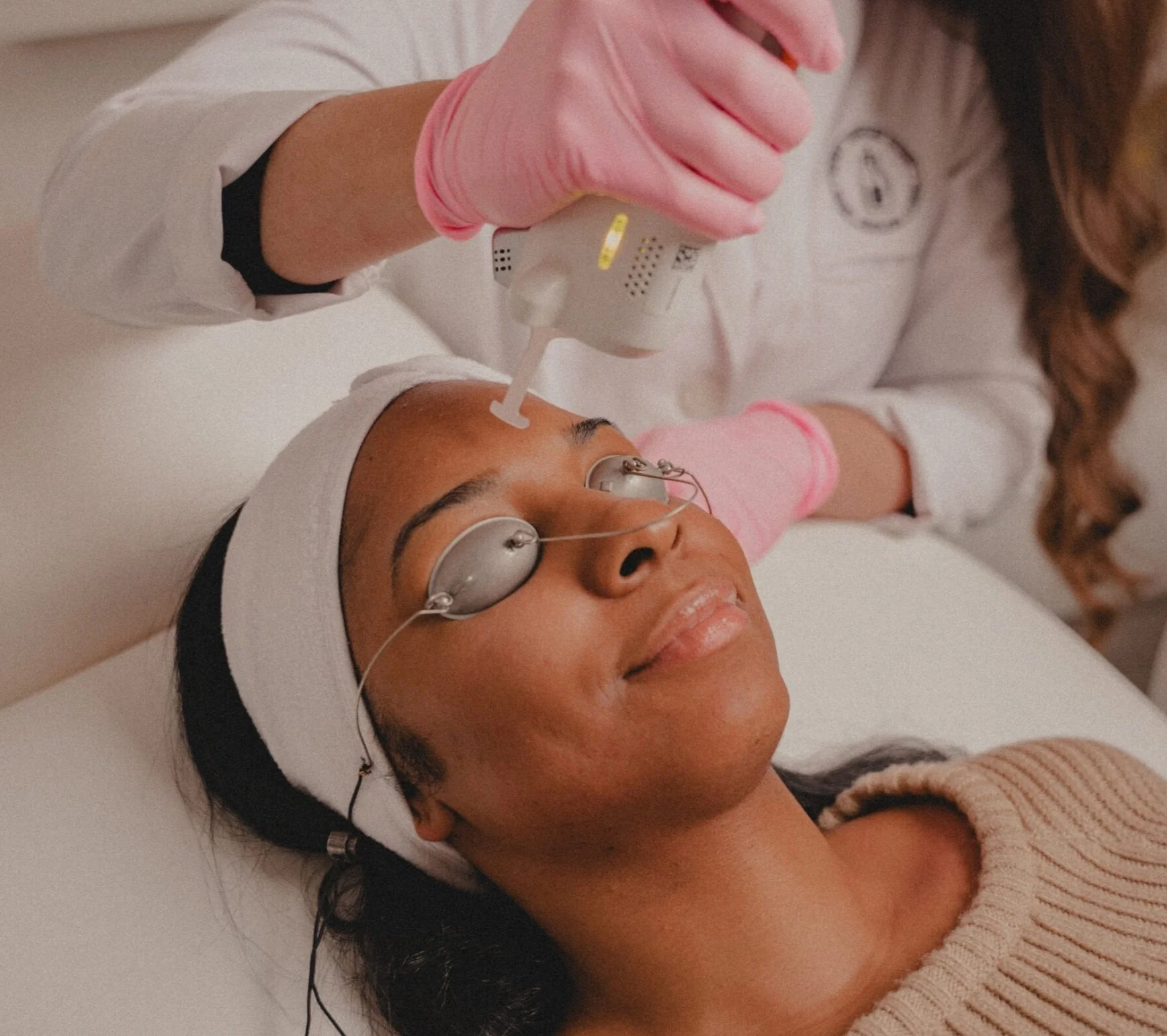 A woman receiving a facial treatment with a derma roller or micro-needling tool while wearing eye patches, lying on a treatment bed with a white headband, and a skincare professional applying treatment with a device.