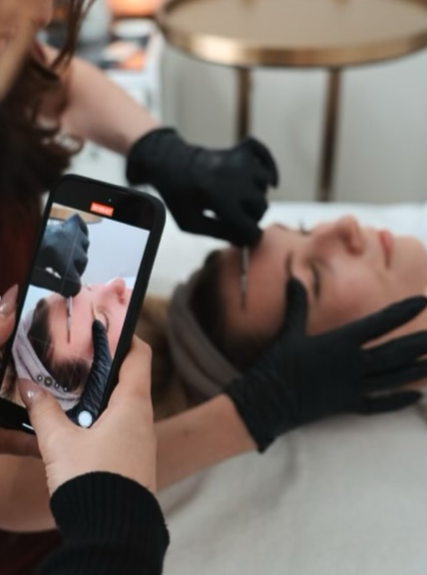 Person taking a close-up photo of a woman lying down with her eyes closed, while a middle-aged woman has makeup or skincare treatment applied with a brush. Both are wearing black gloves, and the scene includes a small round table nearby.