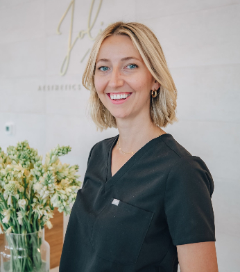 A smiling woman with short blonde hair wearing black scrubs standing inside a clinic or office with a flower arrangement in the background.