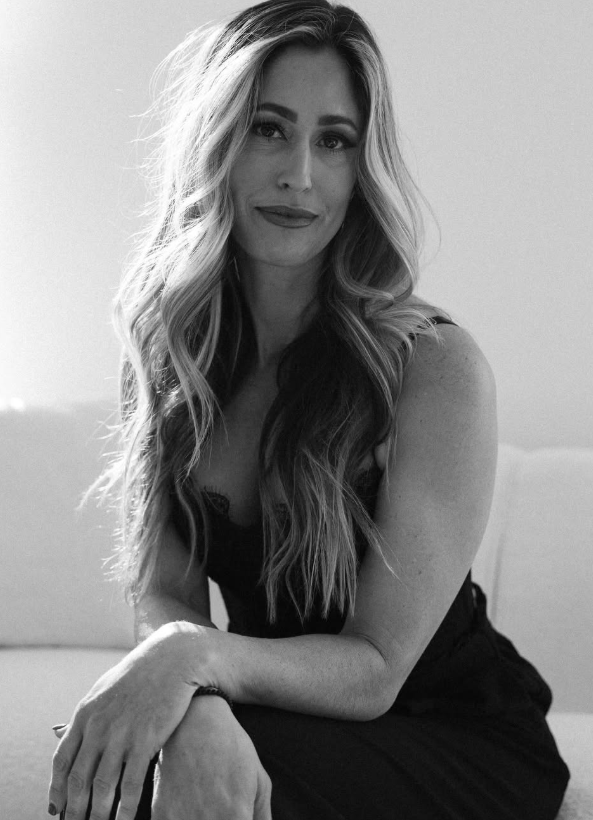 Black and white portrait of a woman with long, wavy hair sitting on a sofa, smiling softly at the camera.