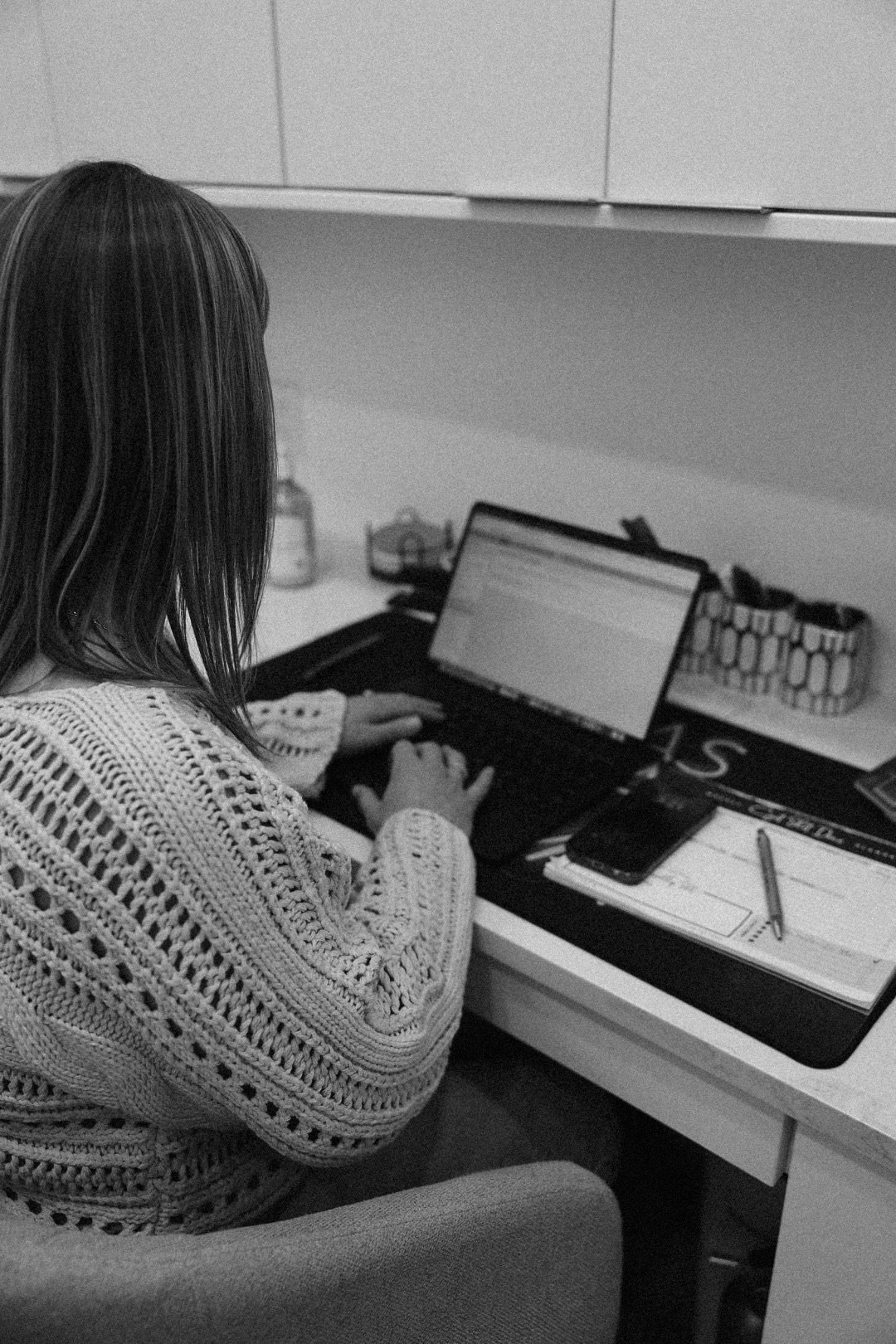 A woman with shoulder-length hair wearing a knitted sweater sitting at a desk working on a laptop, with a notepad, pen, and smartphone on the desk.