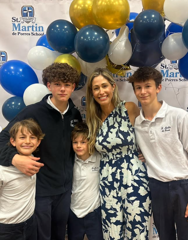 A group of five people, including a woman and four young boys, standing in front of a backdrop with balloons and the logo of St. Martin de Porres School. The balloons are blue, white, gold, and black. The woman is wearing a navy and white patterned dress, and the boys are dressed in white shirts and hoodies.