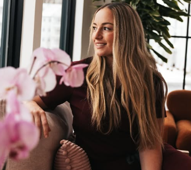 A woman with long blonde hair sitting on a couch in a modern room with large windows and a potted plant, smiling and looking to the side.