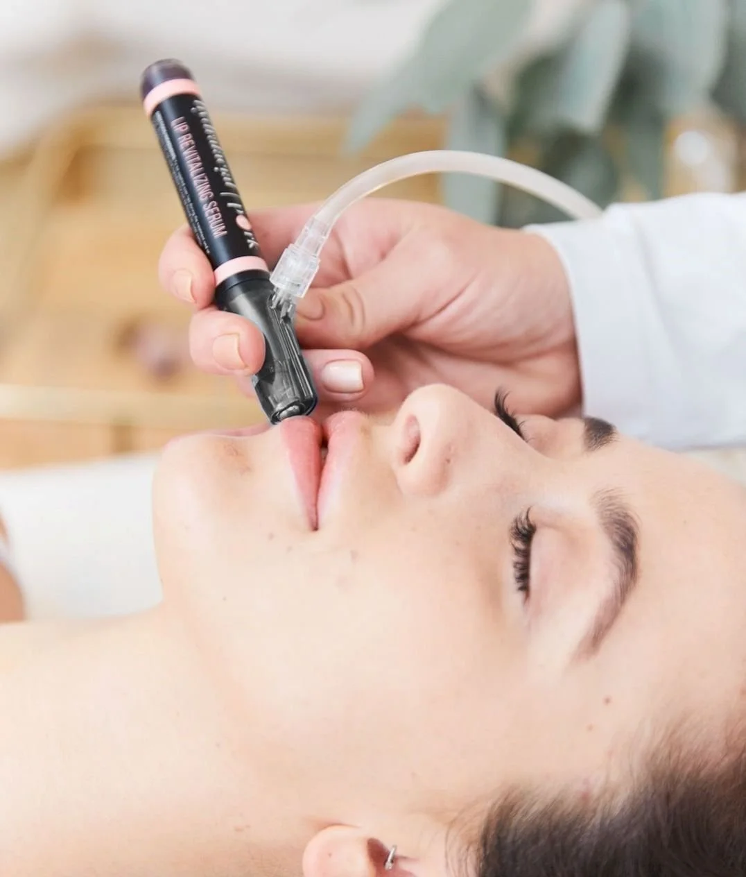 A woman receiving a cosmetic treatment involving a device emitting a pink light applied to her lips by a practitioner.