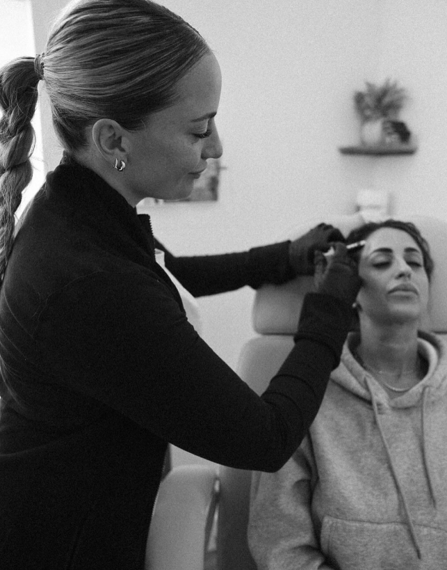 A woman with braided hair, wearing a black jacket and gloves, is applying a cosmetic treatment near the eye of another woman sitting with her eyes closed in a gray hoodie in a clinical setting.