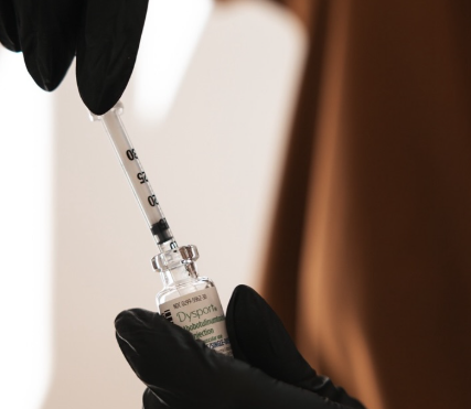 A person wearing black gloves holding a syringe and a small glass vial, likely preparing a vaccine.