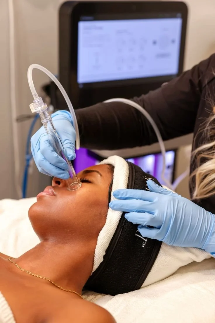 A woman receives a cosmetic injection to her face while lying down in a clinical setting, with a healthcare professional wearing blue gloves administering the treatment.