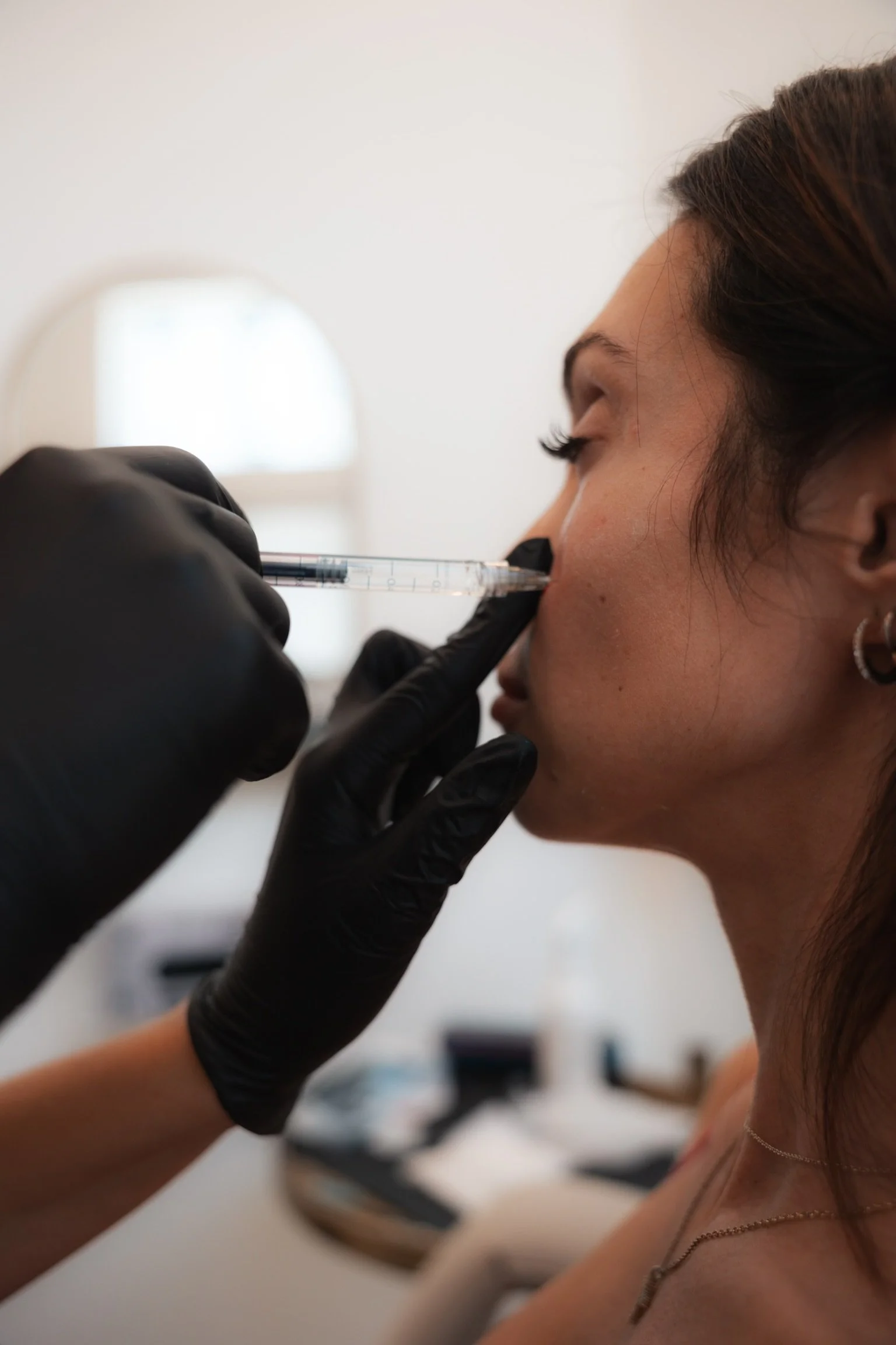 A woman receiving a cosmetic injection in her nose from a healthcare professional wearing black gloves.