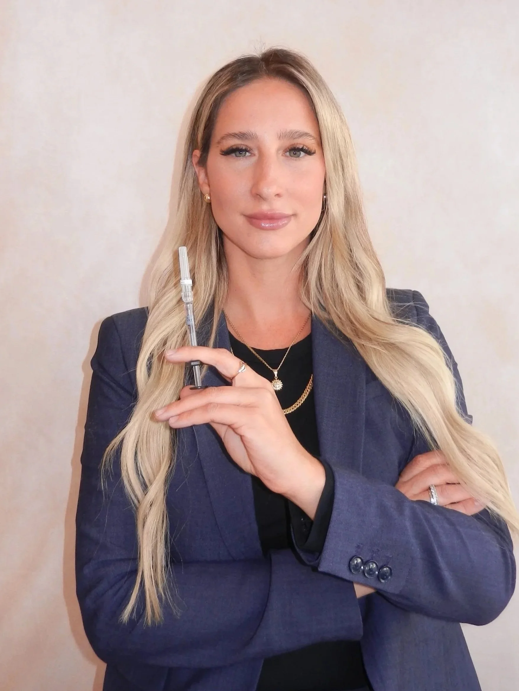 A woman with long blonde hair and blue eyes holding a syringe, dressed in a dark blazer and black top, standing against a light-colored background.