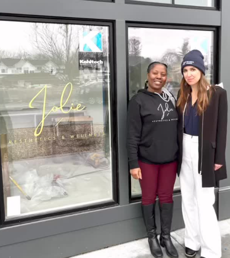 Two women standing outside a storefront with a large glass window, smiling for the photo. One woman is wearing a black hoodie and maroon pants, and the other is dressed in a black coat, white pants, and a navy beanie.