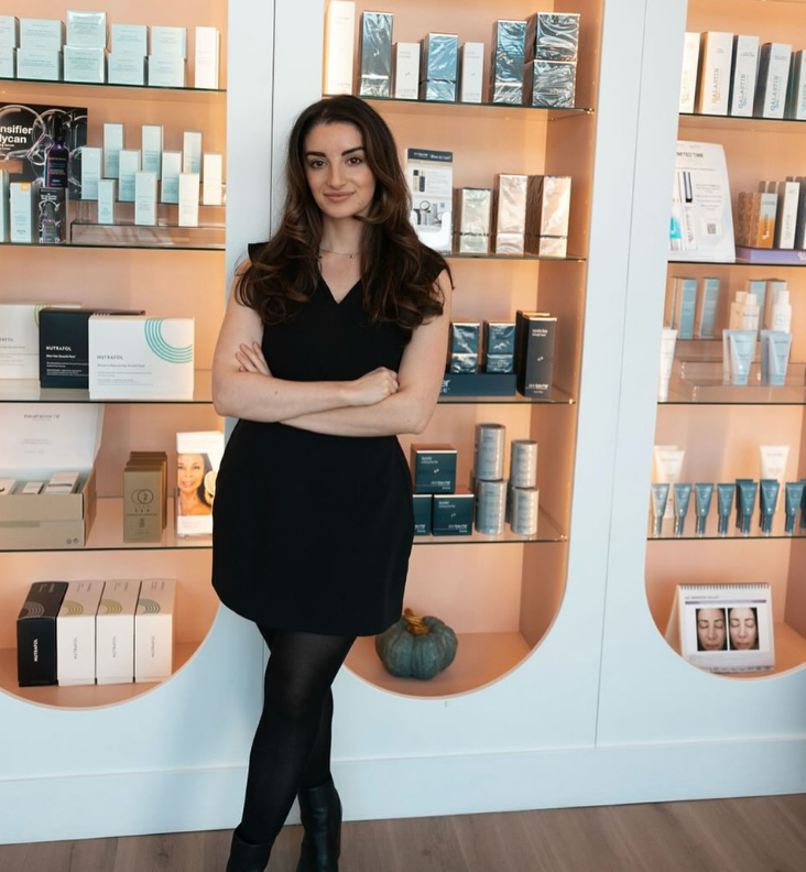 A young woman standing with crossed arms in front of shelves filled with skincare and beauty products.