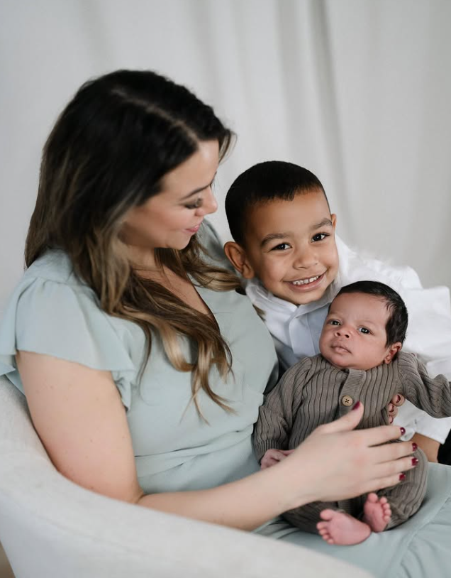 A woman sitting on a couch with her young son and a newborn baby, smiling and looking affectionate.