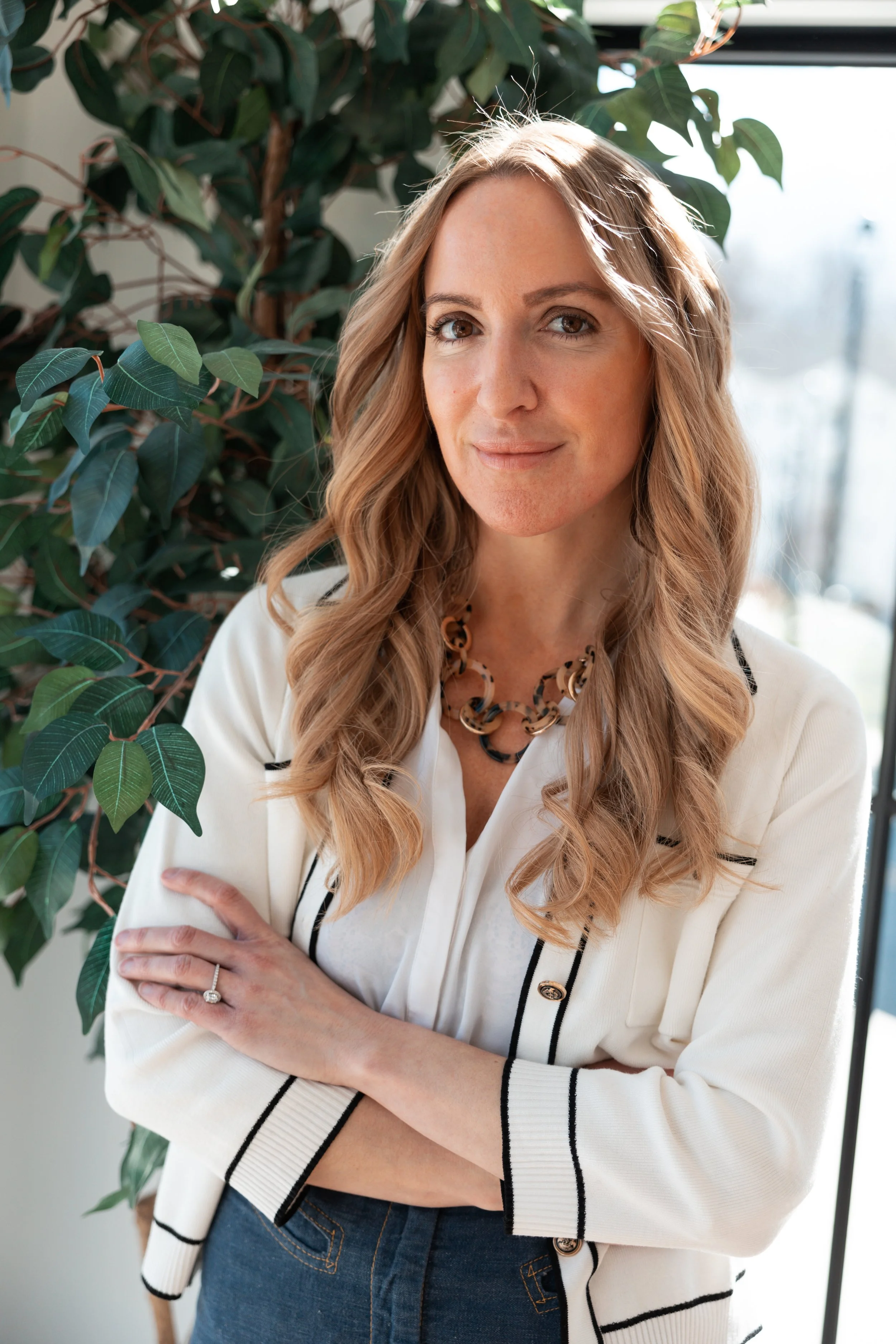 A woman with long, wavy blonde hair standing inside near a large leafy plant, wearing a cream-colored blazer with black accents, a white top, and a chunky necklace, with sunlight streaming in through a window in the background.