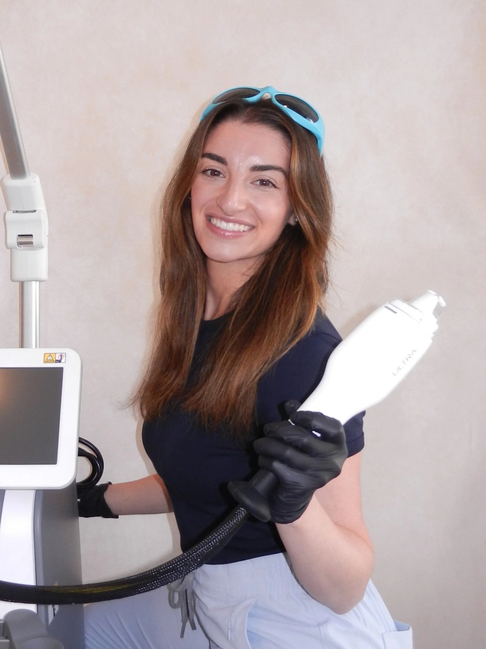 A smiling woman with long brown hair wearing black gloves, blue goggles on her head, and a navy shirt, holding a laser skin treatment device in her right hand.