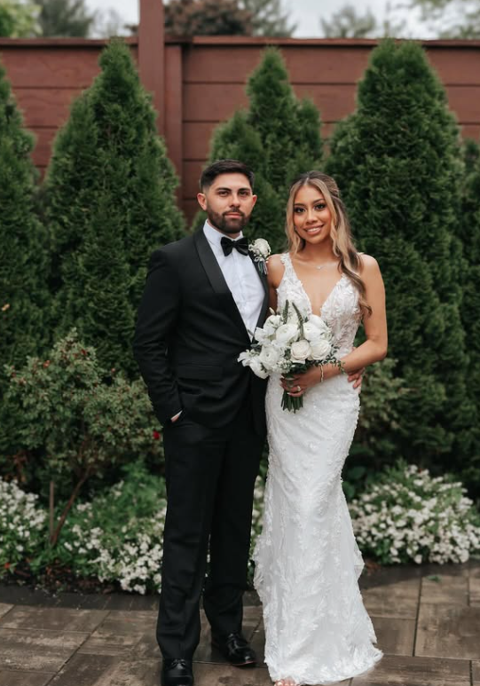 A newlywed couple posing outdoors. The groom is wearing a black tuxedo with a bow tie, and the bride is in a white lace wedding dress holding a bouquet of white flowers. They stand in front of green trees and a wooden fence.