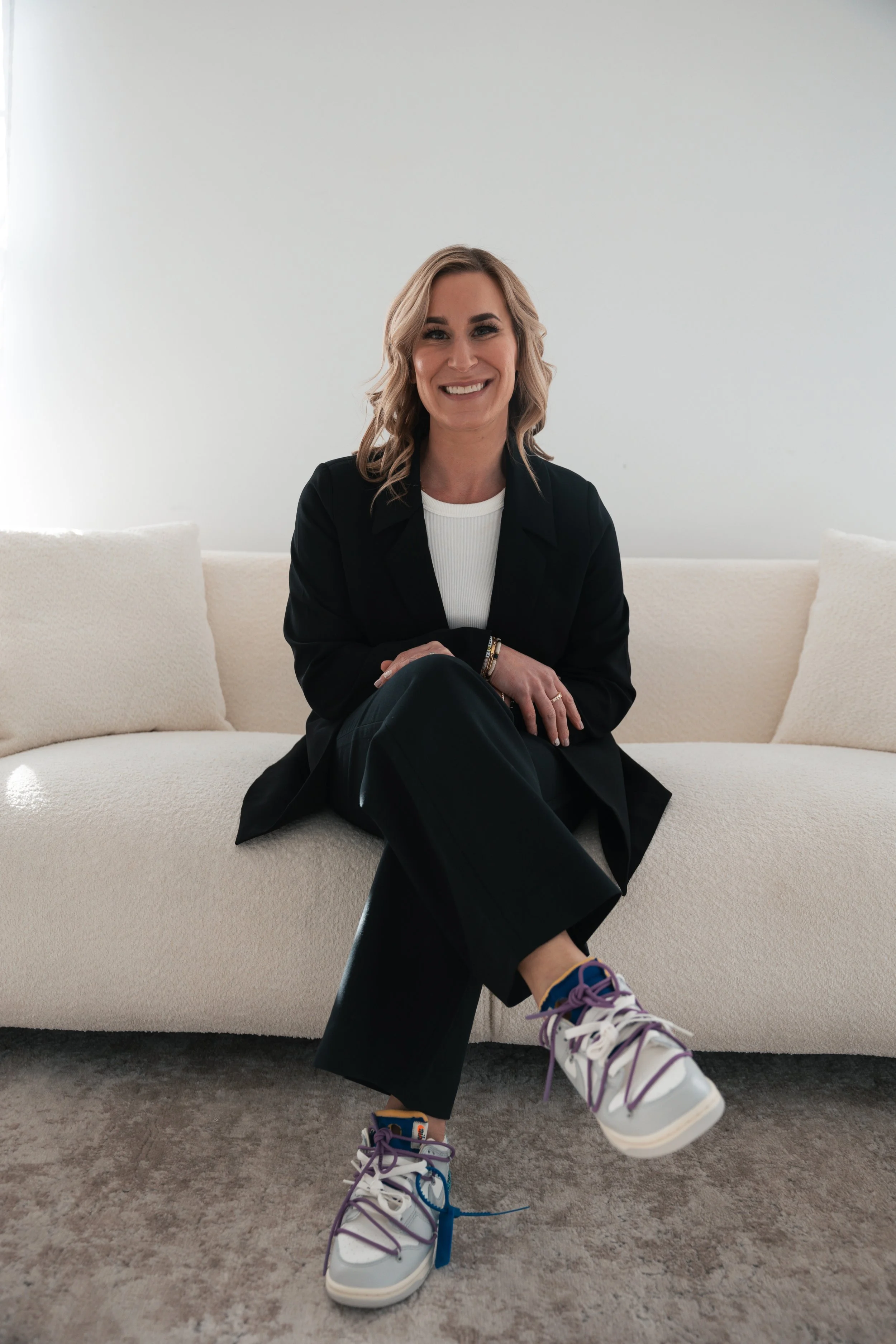 A smiling woman with blonde hair sitting on a white sofa in a minimalistic room, wearing a black blazer, white shirt, black pants, and colorful sneakers.