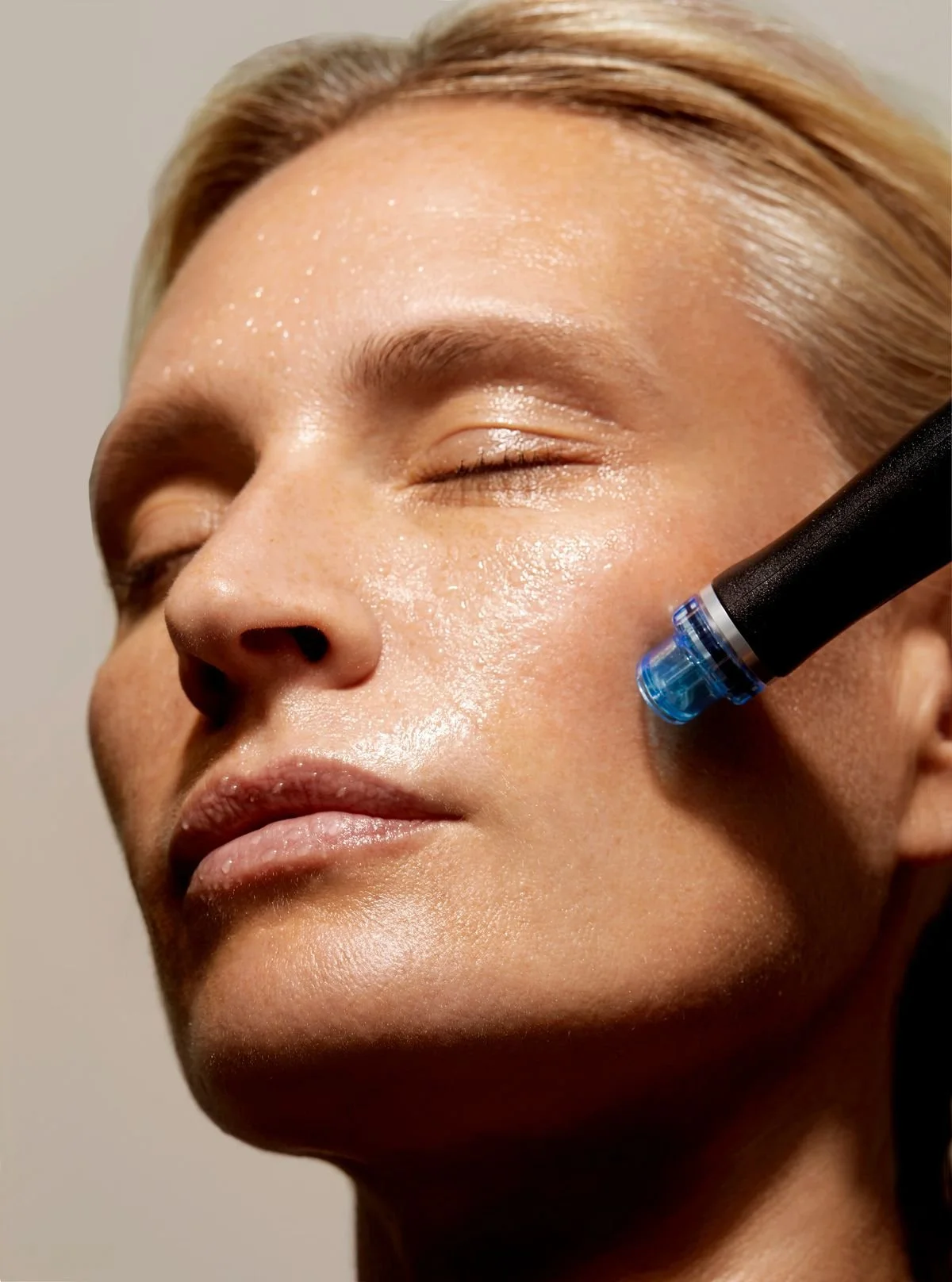 A woman with closed eyes receiving a skincare treatment with a black device applied to her cheek, close-up of her face.