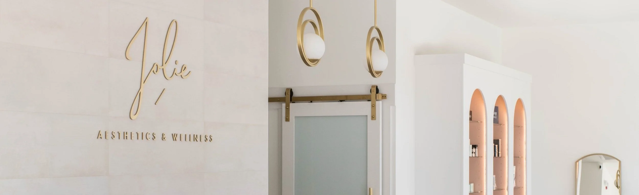 Interior of a modern skincare and wellness clinic with a gold sign that reads 'jolie Aesthetics & Wellness', gold hanging lights, a sliding door, and built-in shelves with skincare products.