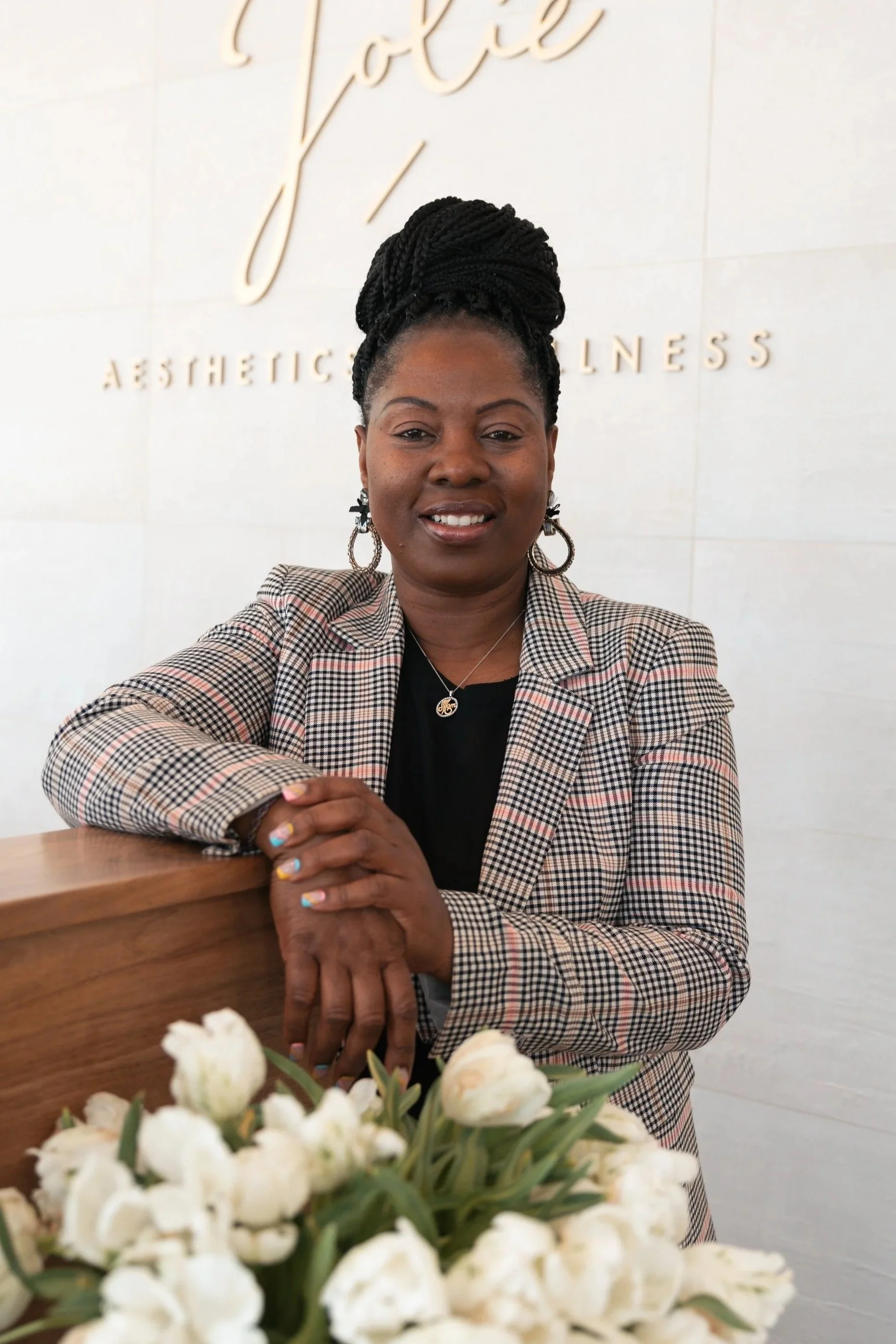 A woman with braided hair wearing a plaid blazer and jewelry, standing behind a wooden counter with white flowers, inside a wellness center with a sign saying 'Aesthetic Wellness' in the background.