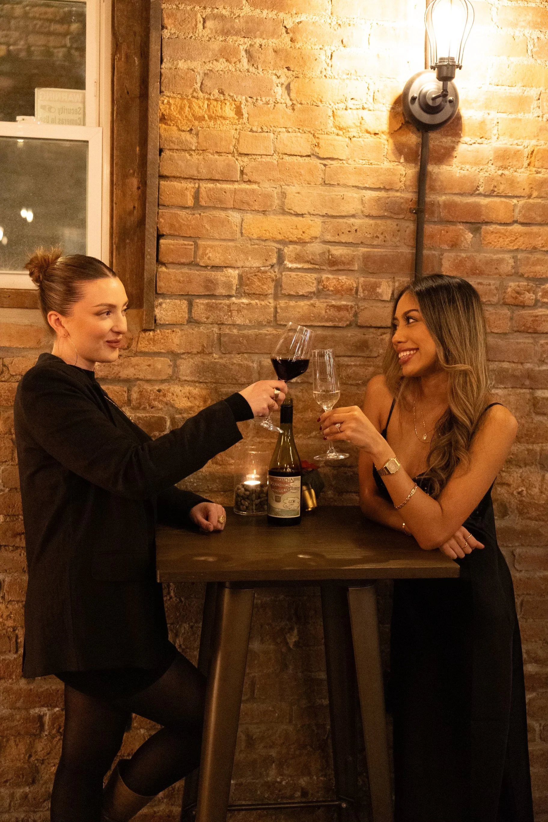 Two women clinking wine glasses at a high table in a cozy, brick-walled restaurant or bar.