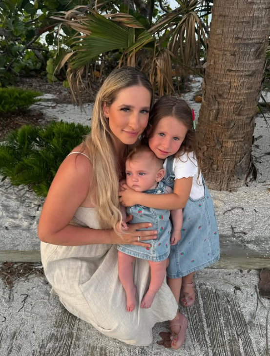 A woman and two young girls sitting on a wooden bench at the beach. The woman has long blonde hair, wearing a light-colored dress, and is smiling at the camera. The older girl, with red hair and a flower hair clip, is hugging the younger girl, who has light brown hair and is wearing a denim dress. They are surrounded by sand, palm trees, and tropical plants.