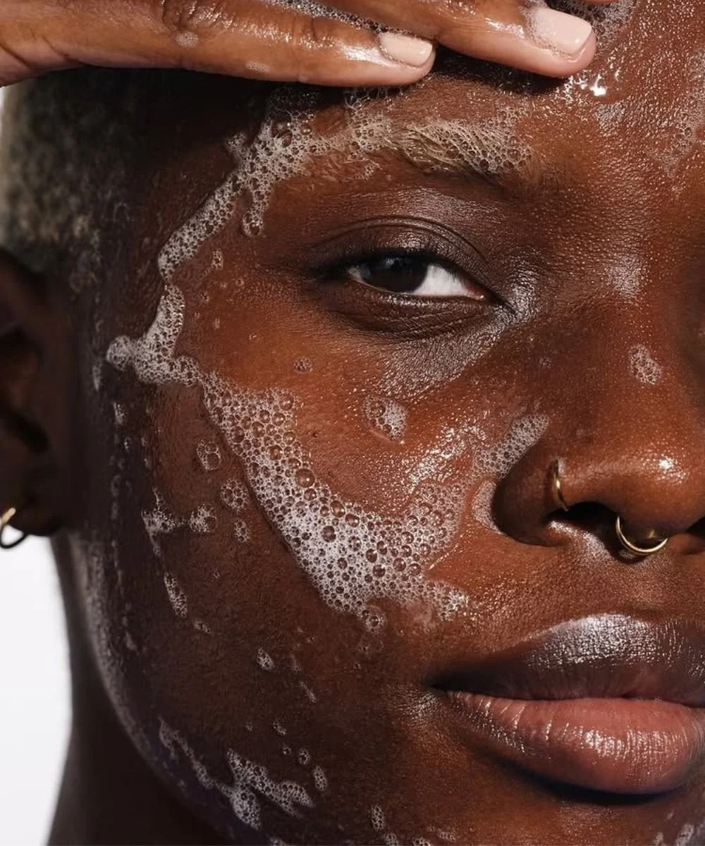 A person with dark skin washing their face with soap, creating foam on their face.