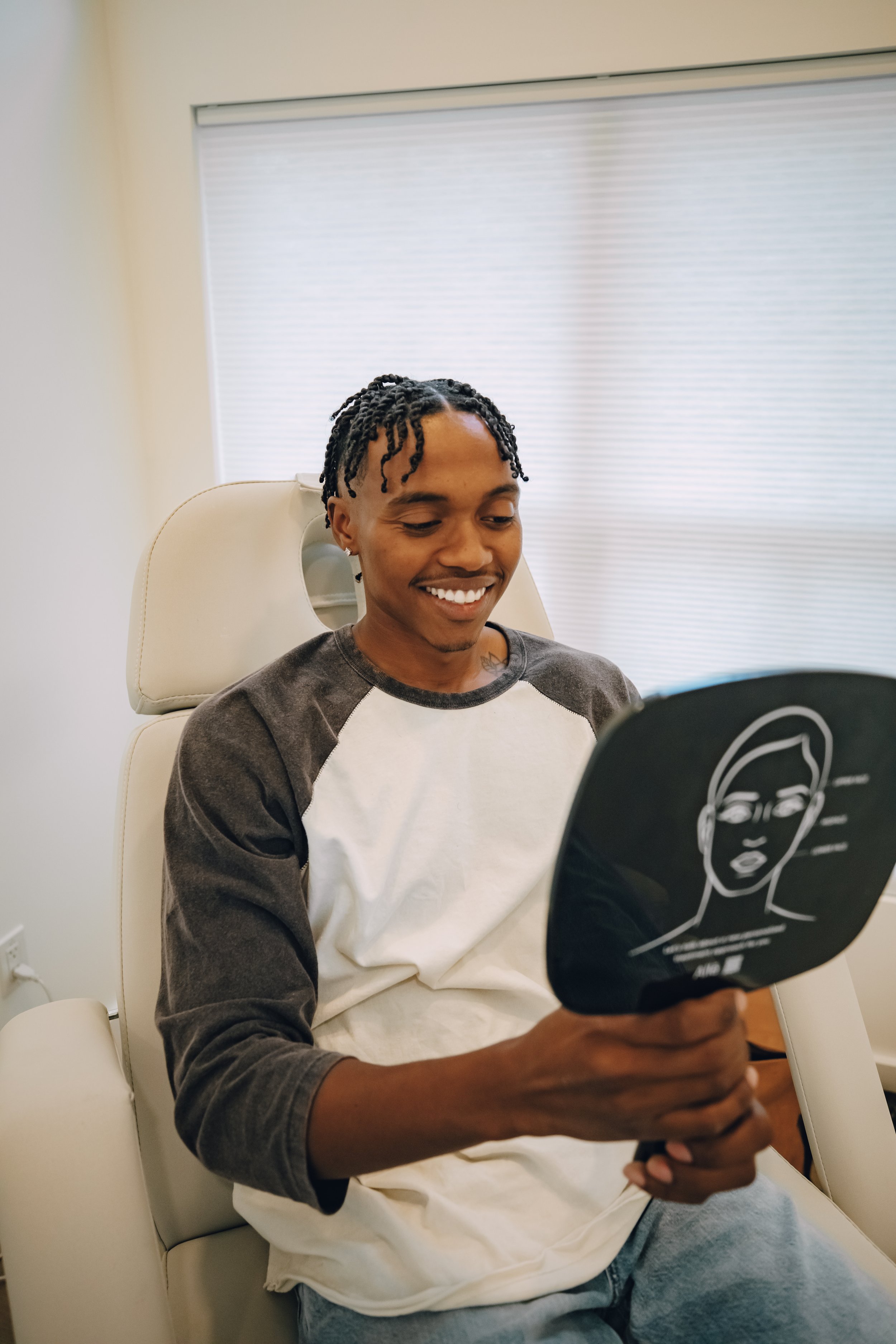 Young man sitting in a medical chair, smiling while holding a handheld mirror to examine his face.