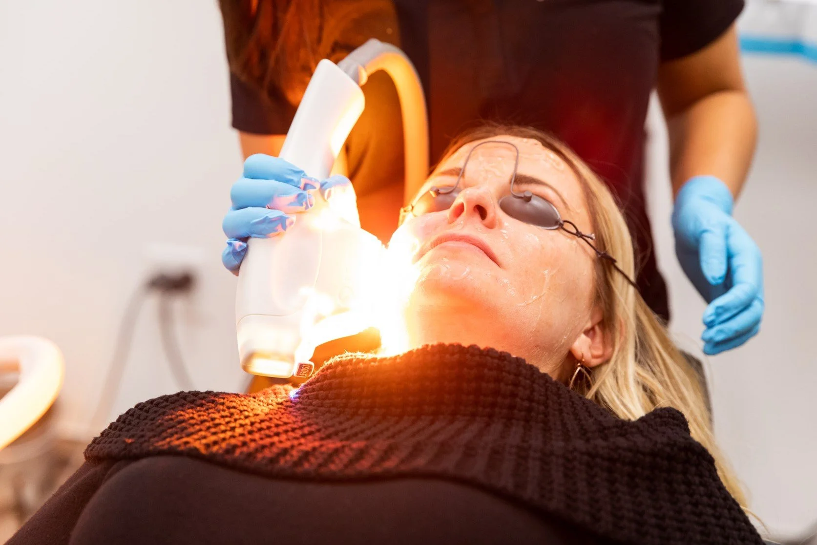 A woman undergoing dental treatment is lying back with a dental device over her eyes. A dental professional wearing blue gloves is holding a curing light to her mouth, illuminating her teeth.