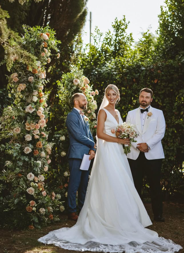 A wedding ceremony outdoors with a bride in a white gown holding a bouquet, a groom in a white tuxedo jacket, and a man in a blue suit standing beside them under a floral arch made of roses and greenery, with trees and sunlight in the background.
