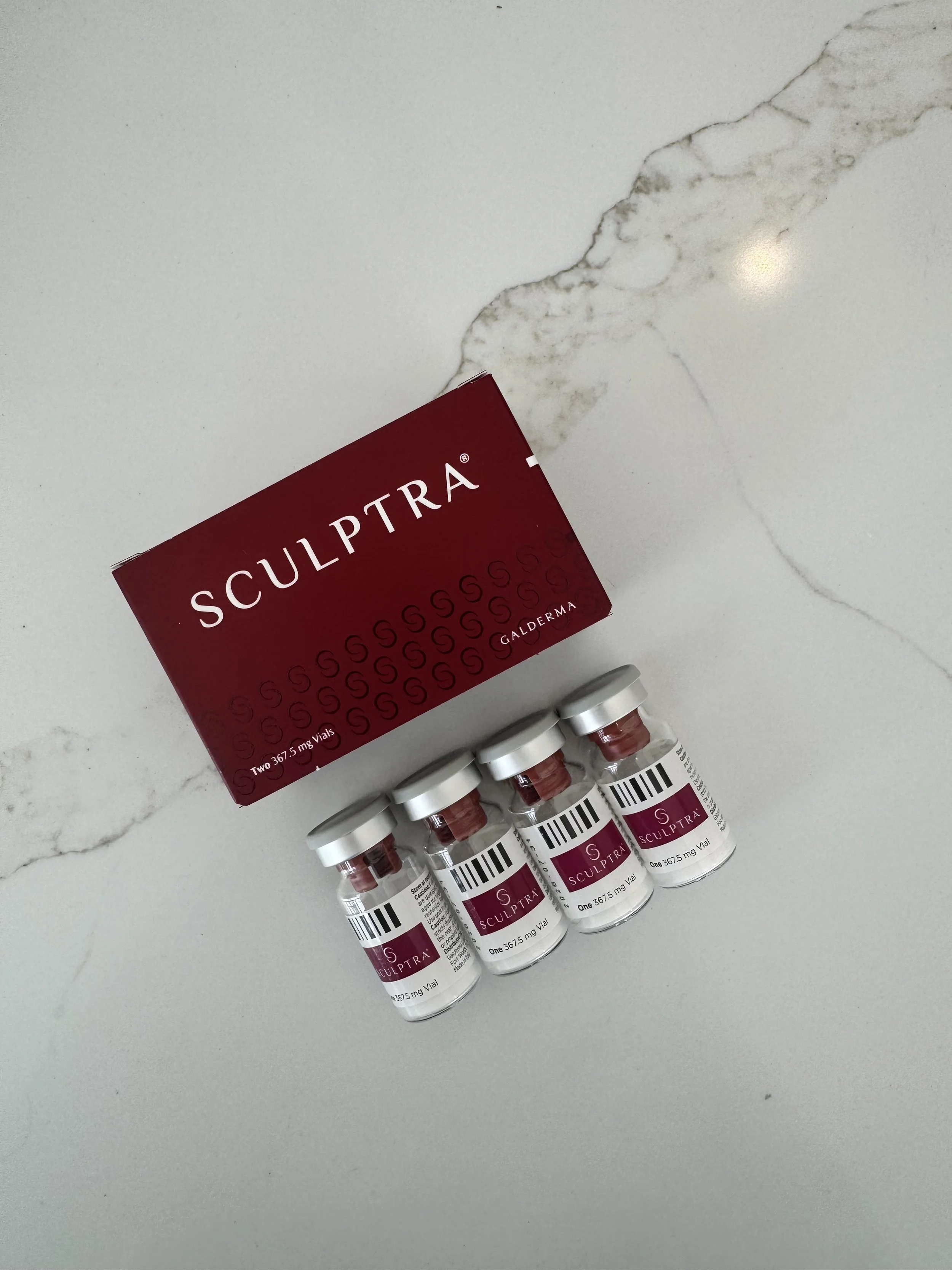 Four vials of Sculptra Aesthetic, a dermal filler, laid on a white marble surface beside its red packaging box.