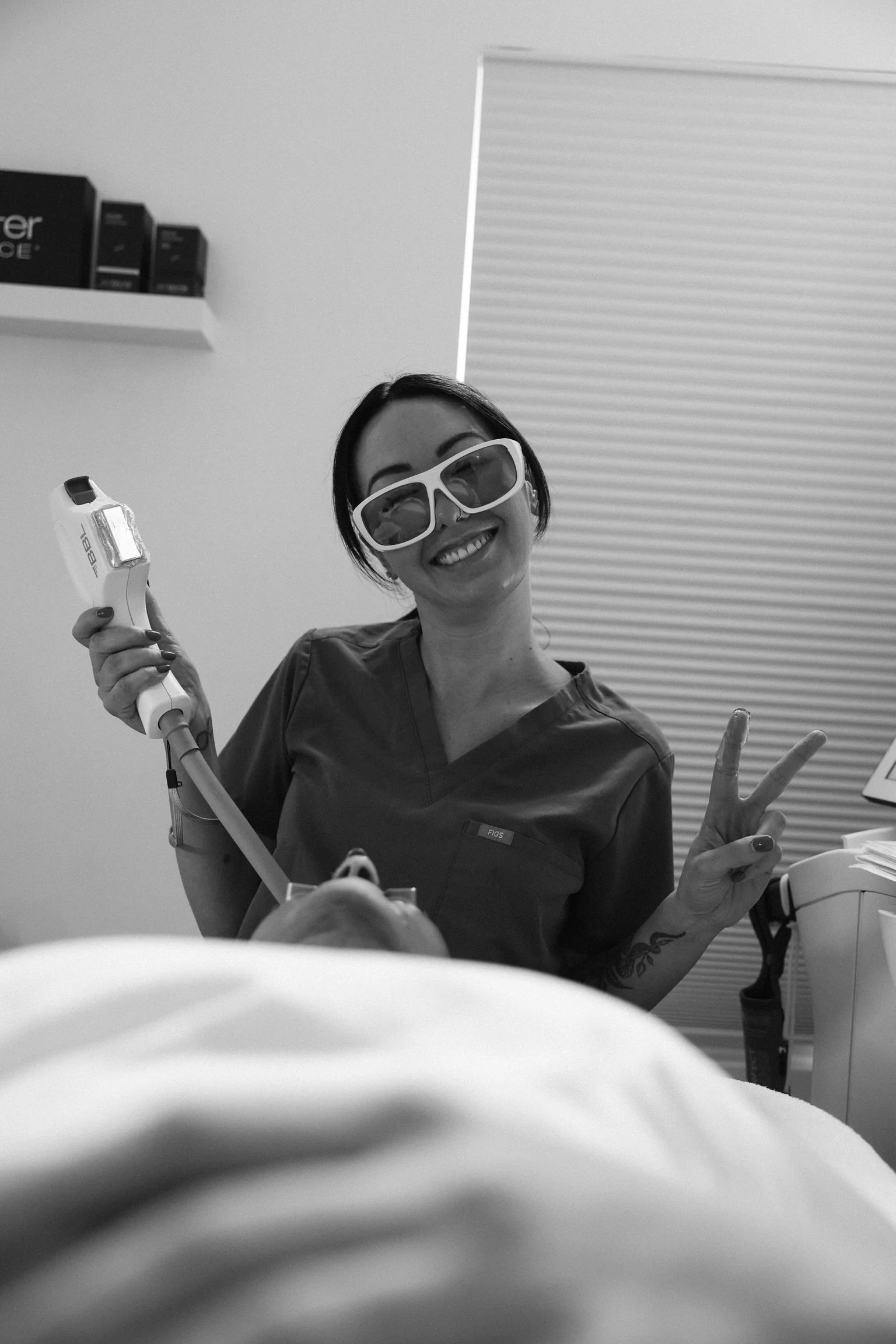 A woman in medical scrubs wearing sunglasses, smiling and making a peace sign, holding a medical device in a clinical setting, with a patient lying in the foreground.