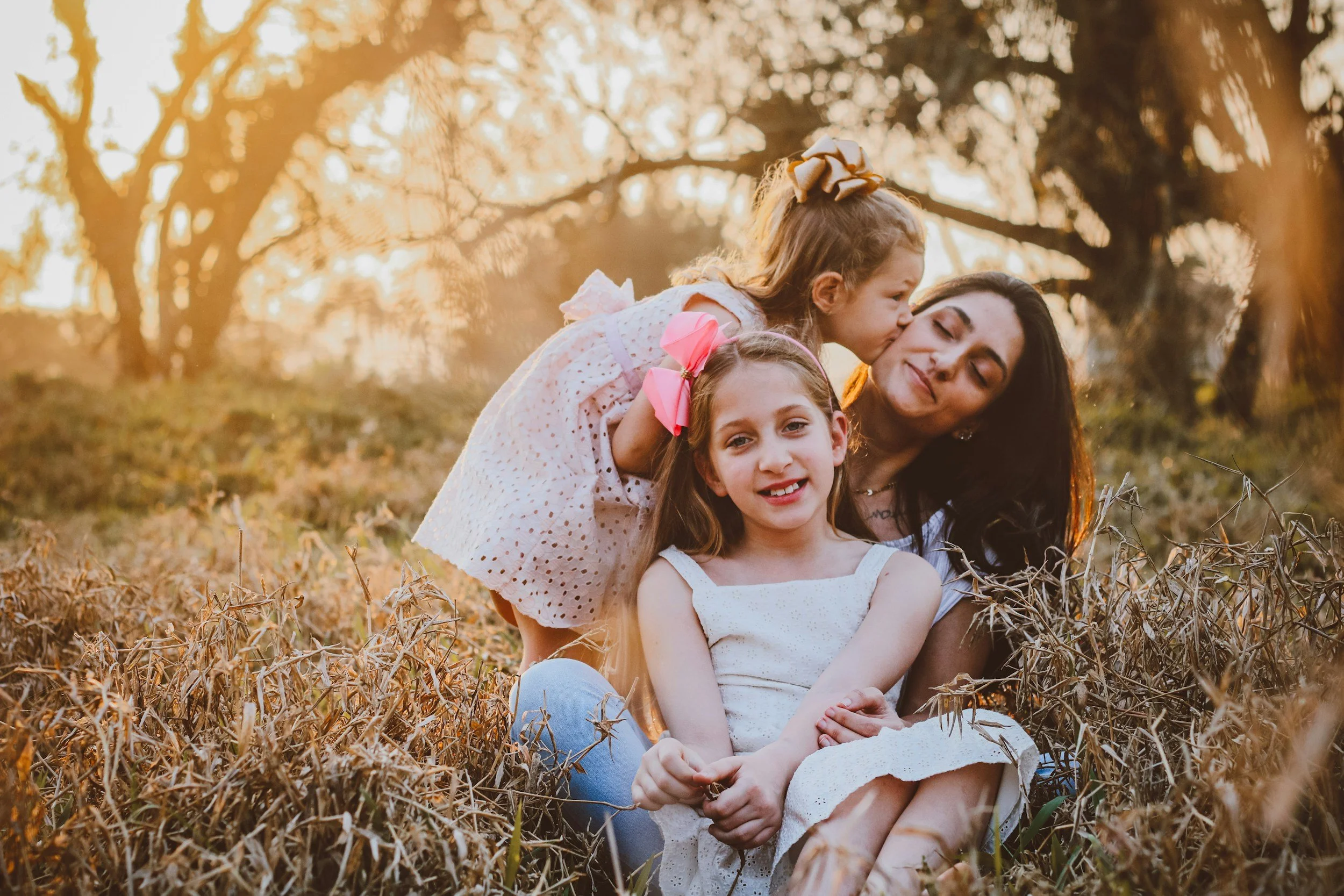 Mother sitting in grassy wooded area with two daughters, one on her lap and the other kissing her cheek. The mother is leaning away from the kiss.