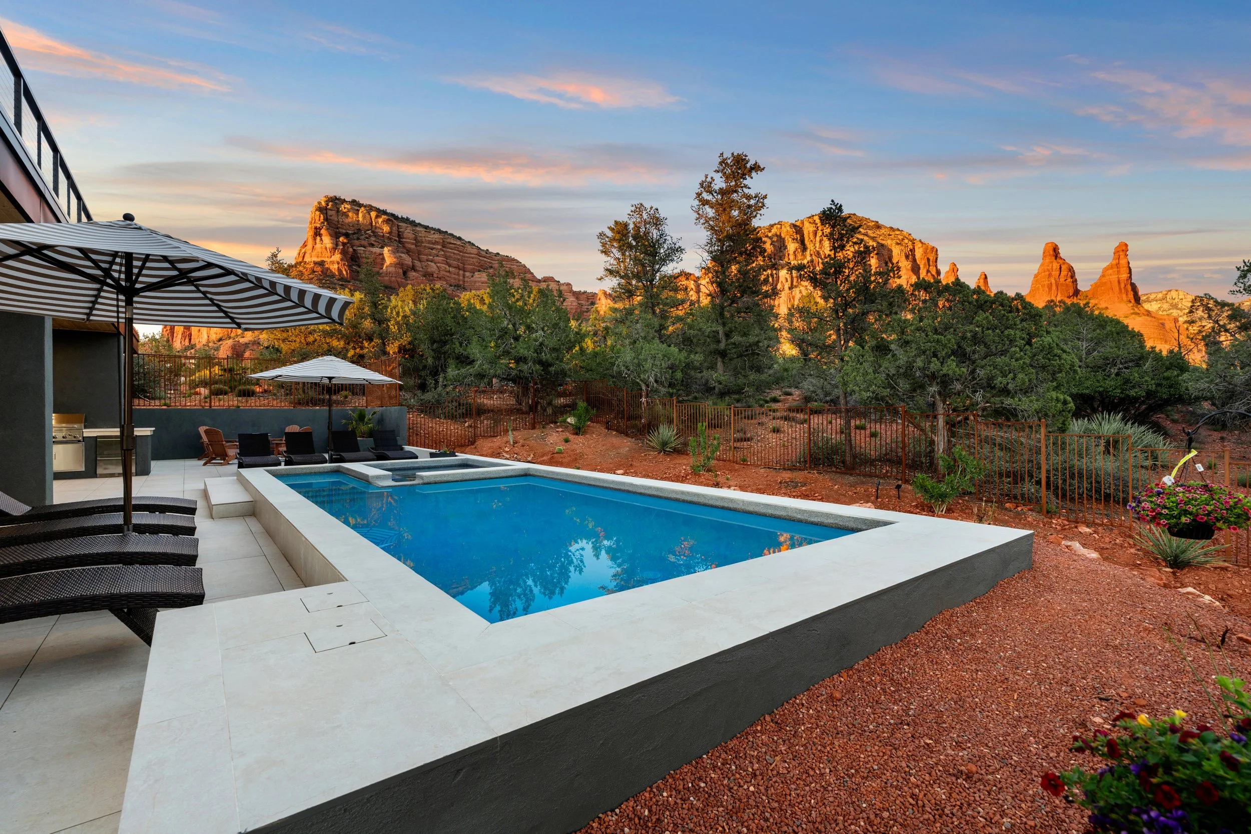 View of a modern outdoor pool area with lounge chairs and umbrellas, set against a backdrop of red rock formations and a partly cloudy sky at sunset in Sedona, AZ