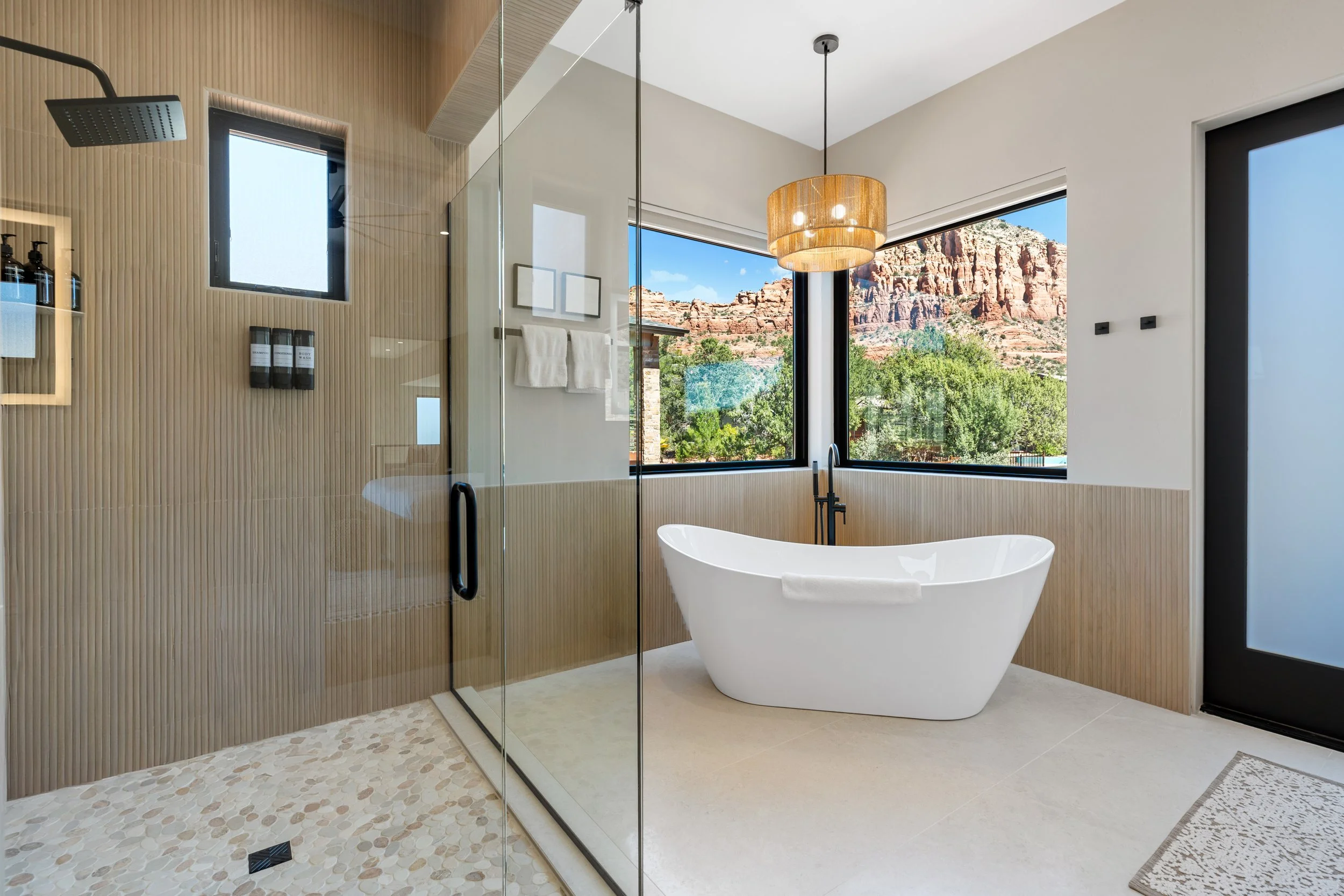 Modern bathroom with a white bathtub, a glass-enclosed shower, large windows showing a red rock landscape, a hanging light fixture, and beige and wood finishes. Luxury home in Sedona, AZ