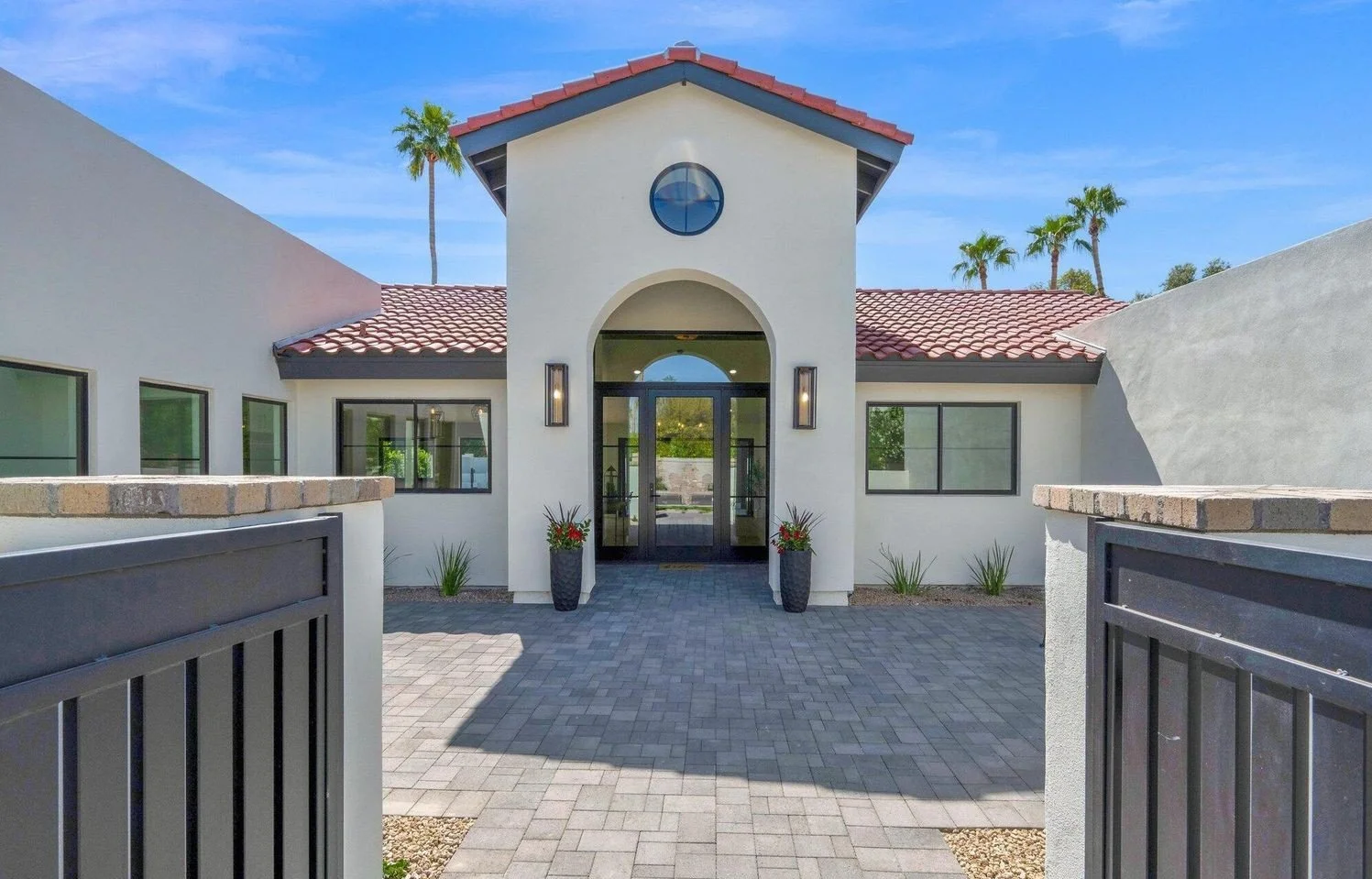 Front view of a modern white house with a red tile roof, black framed windows, and a central arched entrance, and a black gate in the foreground under a blue sky with palm trees in the background. Modern Hacienda, luxury home. Scottsdale, AZ