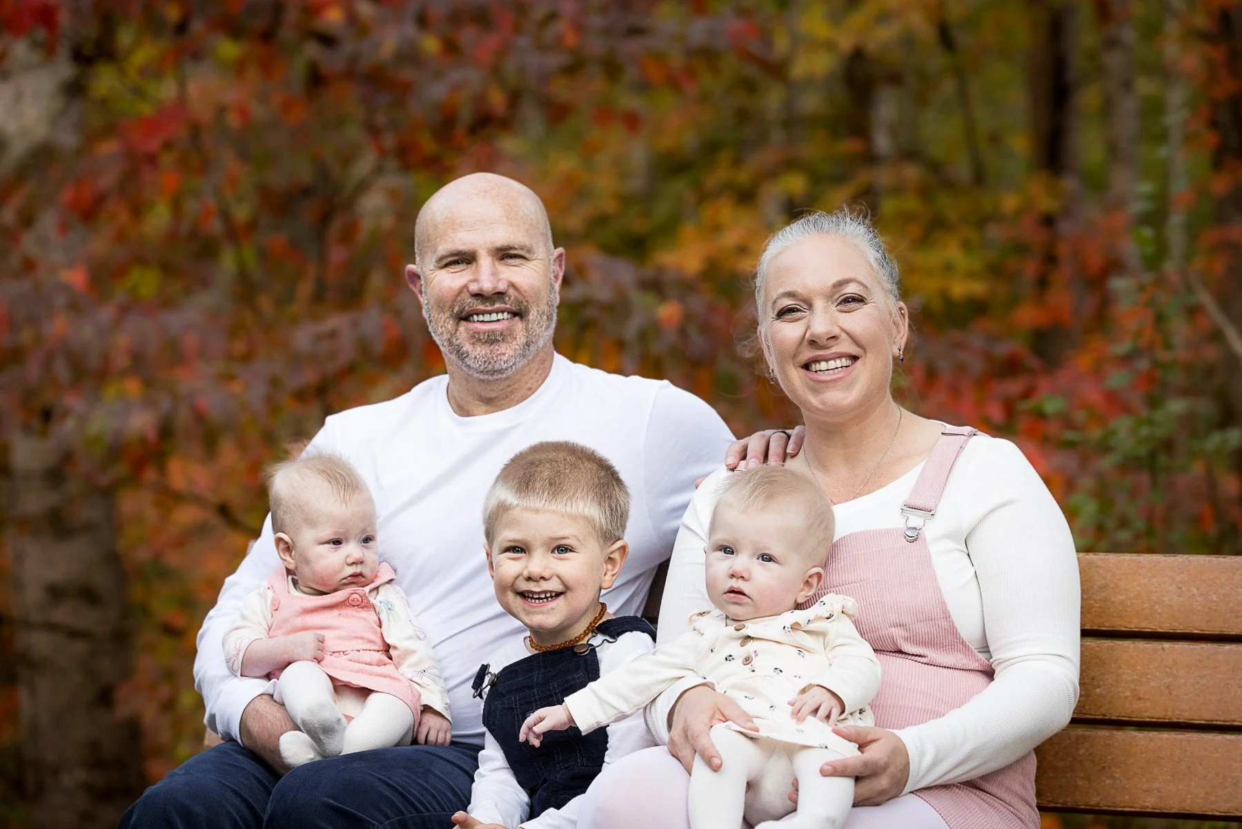 Family of six sitting on a park bench during autumn, including a smiling older man and woman with four young children, surrounded by red and orange fall foliage.