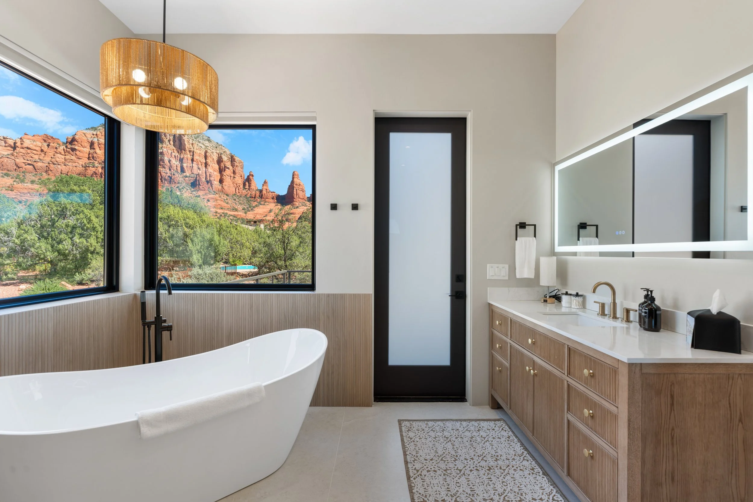Modern bathroom with a freestanding bathtub, large window showing red rock formations and mountains, wooden vanity with gold knobs, illuminated mirror, black door, and neutral decor. Luxury home in Sedona, AZ