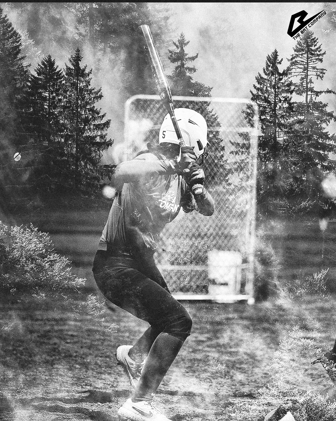 A person wearing a helmet and sports gear holding a baseball bat in a ready stance, with a forest and trees in the background.