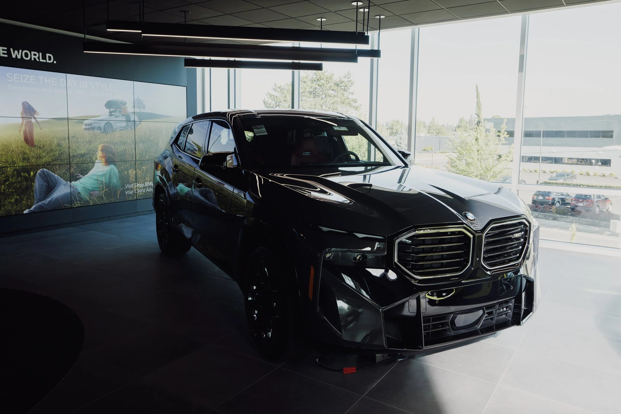 Black luxury car on display inside a modern dealership with large windows and a digital billboard showing outdoor scenery and people.