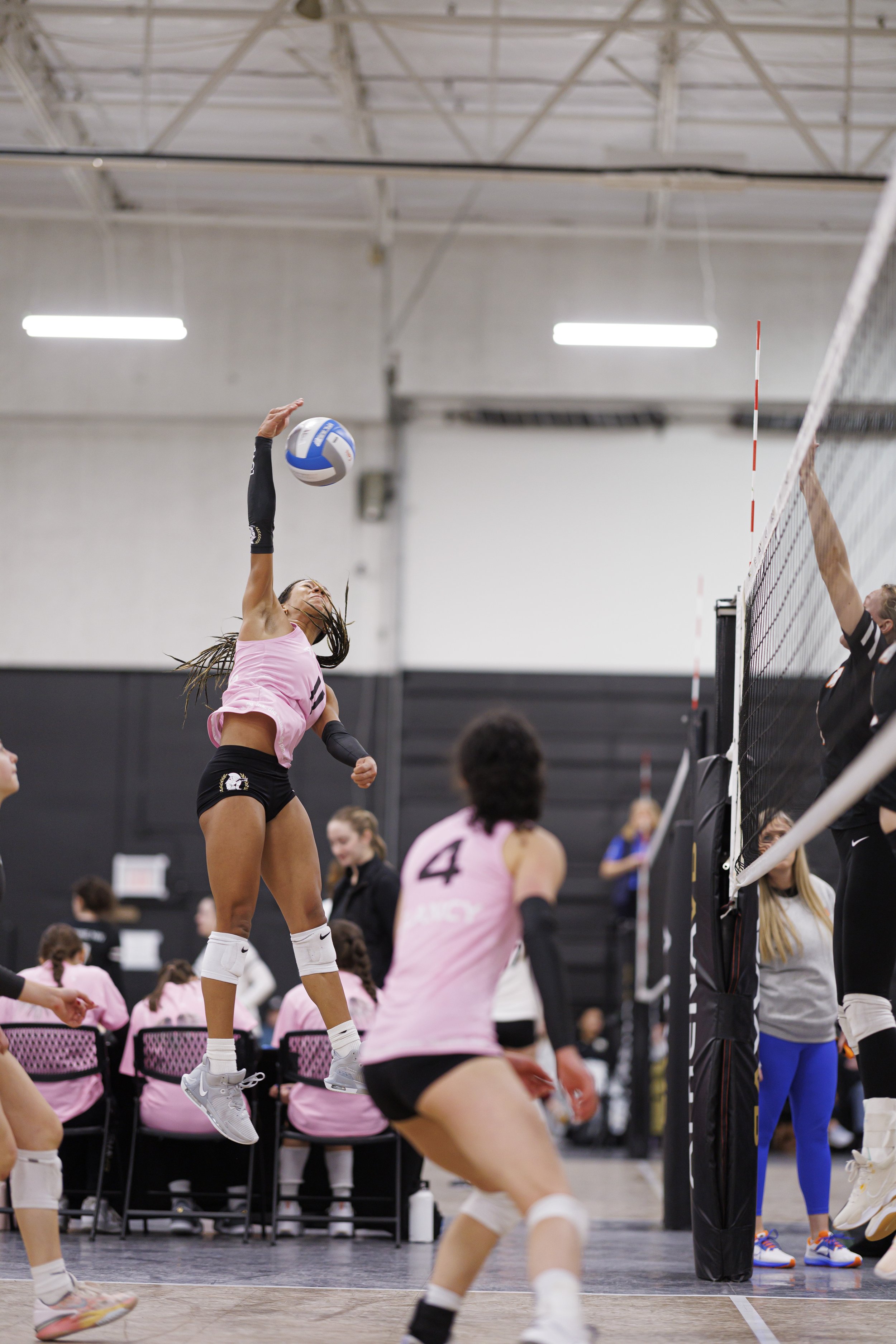 A volleyball game in progress with players in pink jerseys; a player is jumping to spike the ball, while others are preparing to defend. The setting is an indoor sports facility with spectators and officials present.
