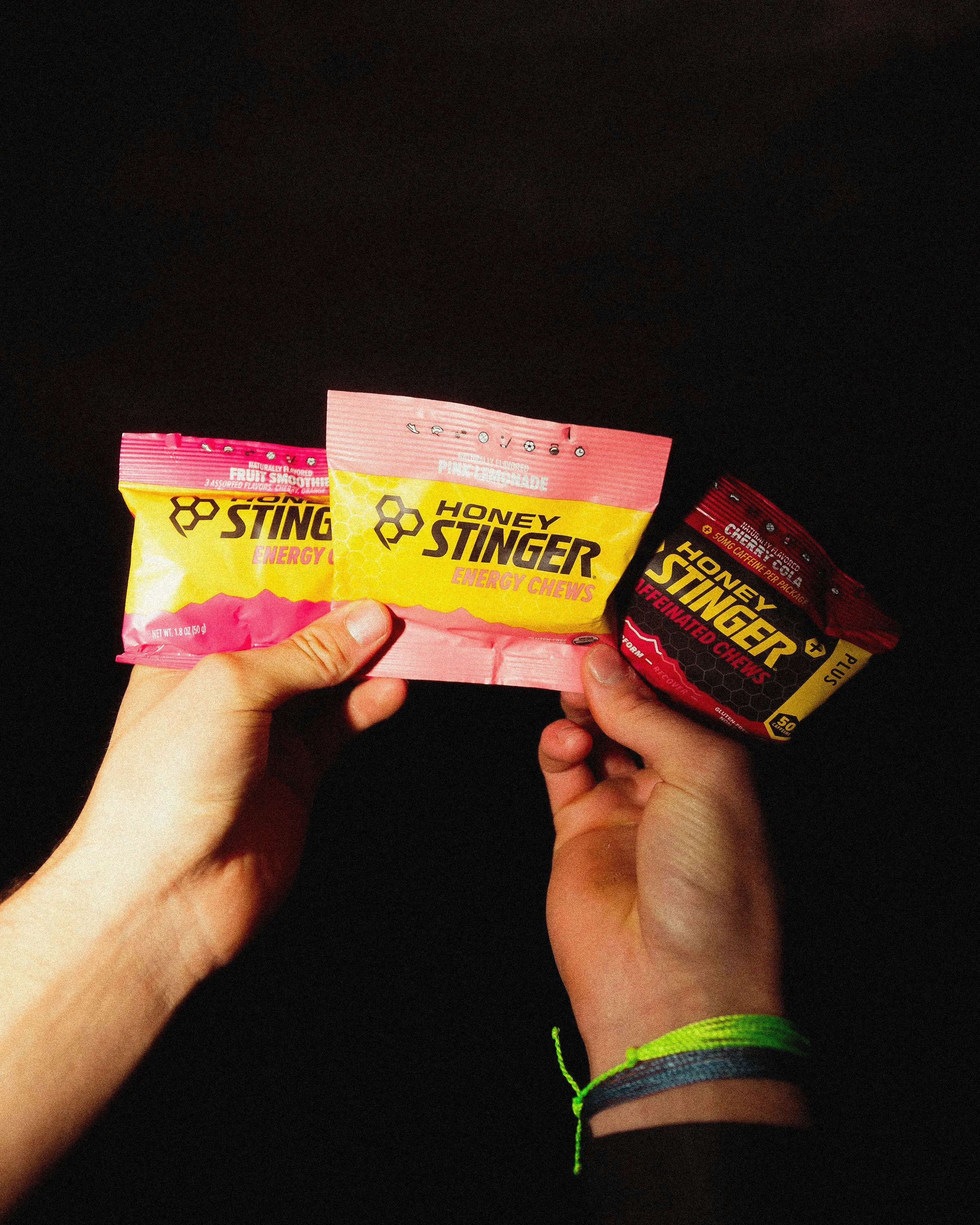 Three packets of Honey Stinger energy chews in pink, yellow, and red packaging, held by two hands with a black background.